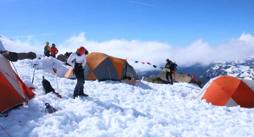 High on a mountainous landscape, tents rest in the snow of a campsite. People are standing around wearing cold-weather gear.